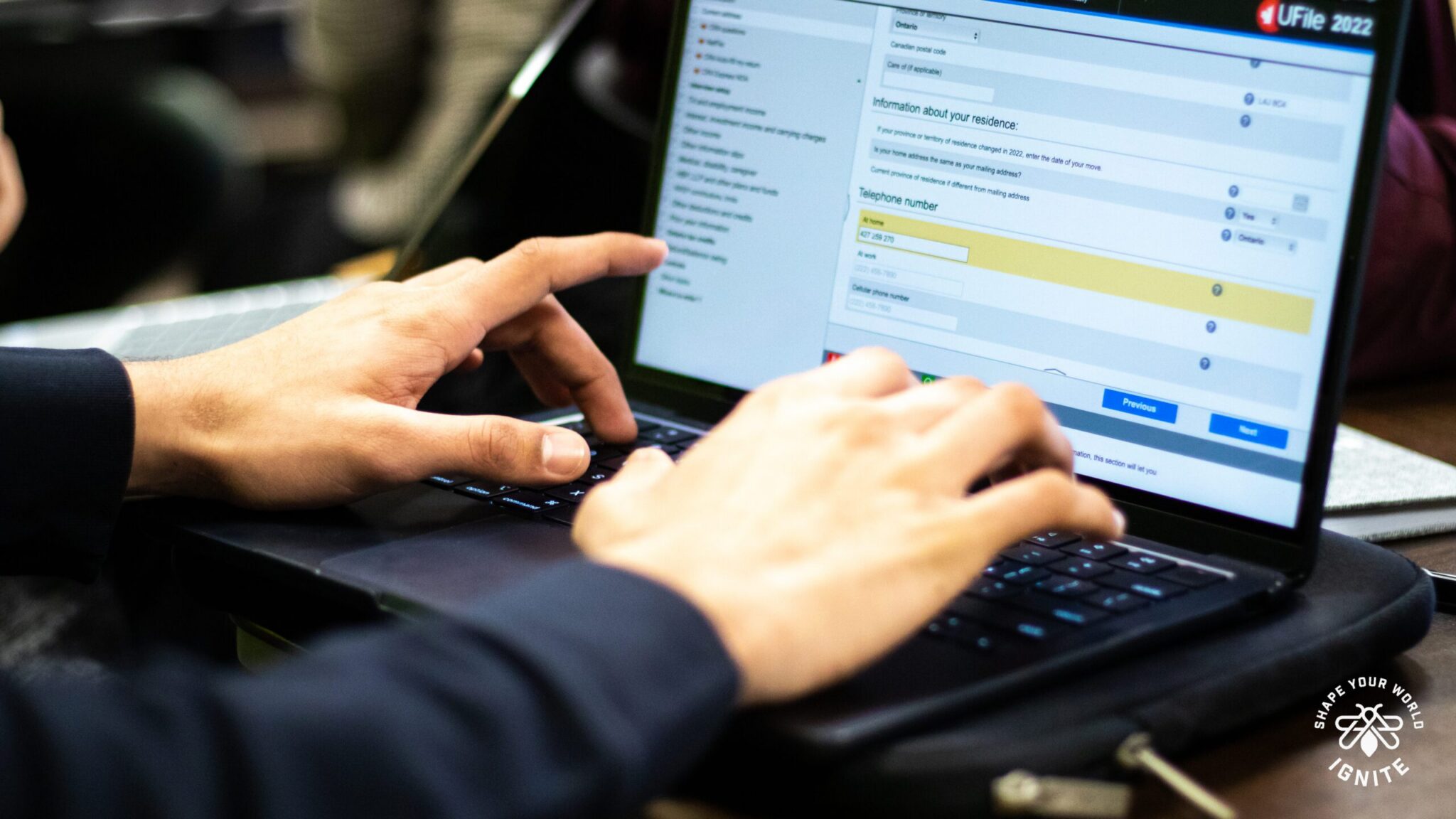 A person types on a laptop, filling out an online form with fields for address and telephone number visible on the screen. The computer rests on a table with other items nearby.