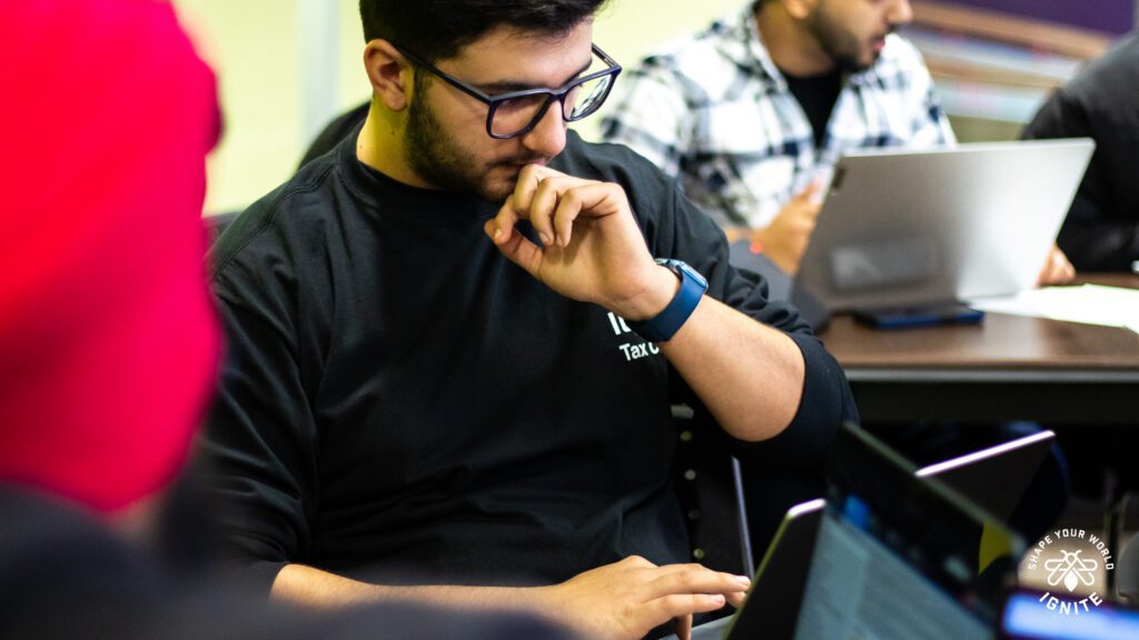 A man wearing glasses and a black shirt is sitting at a desk, looking thoughtfully at a laptop. Other people with laptops are visible in the background, engaged in work or discussion.
