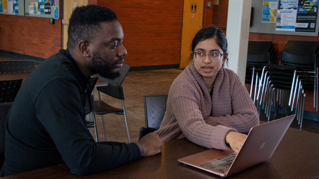 Two people sit at a table indoors, looking at a laptop. The woman gestures toward the screen while the man listens attentively. They appear to be discussing something on the laptop in a casual, public space.