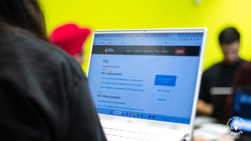 A person using a white Dell laptop, browsing a FAQ page on the ULife website; blurred people and a bright yellow-green wall are in the background.