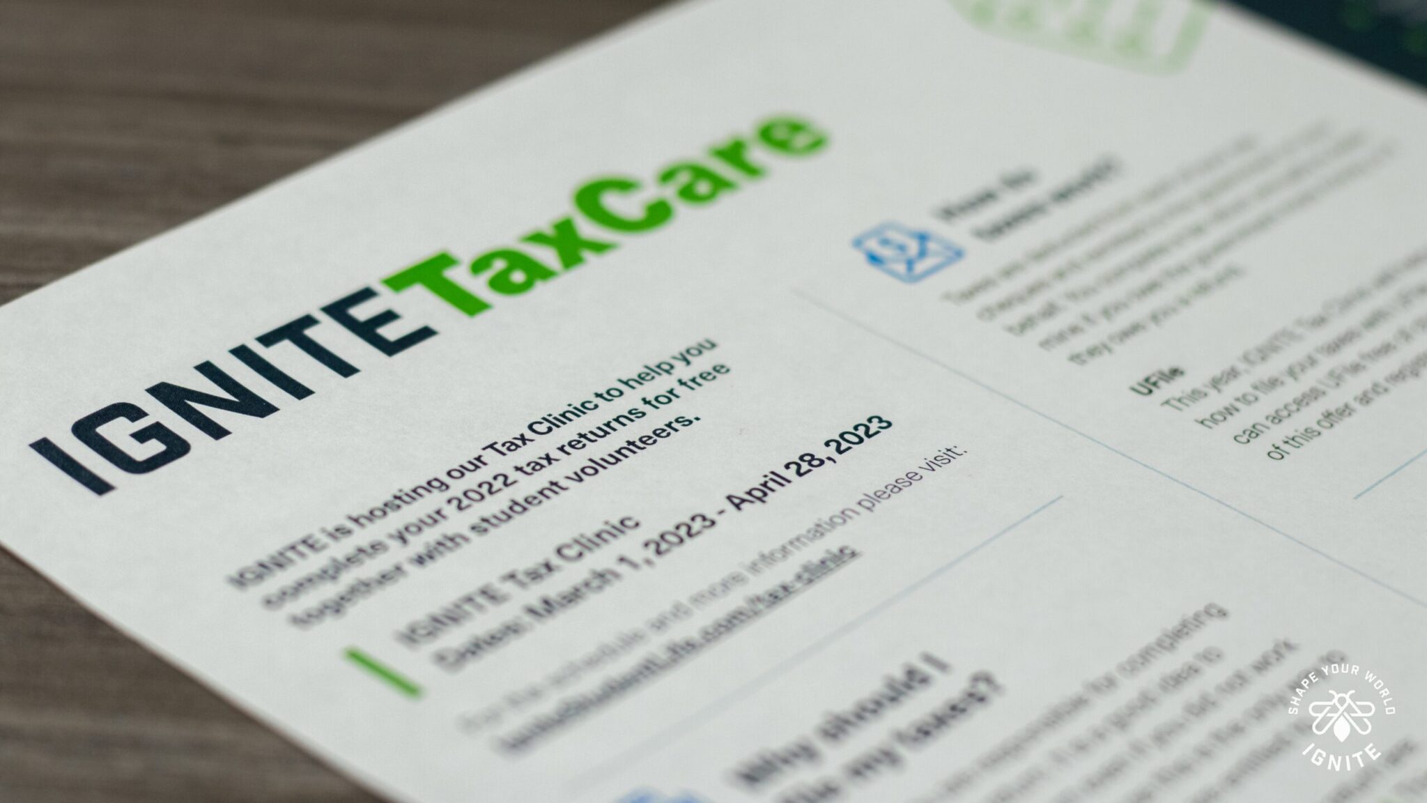 A close-up of a printed flyer titled IGNITE TaxCare, advertising a free tax clinic for 2022 tax returns, with event dates and details partially visible, lying on a wooden surface.