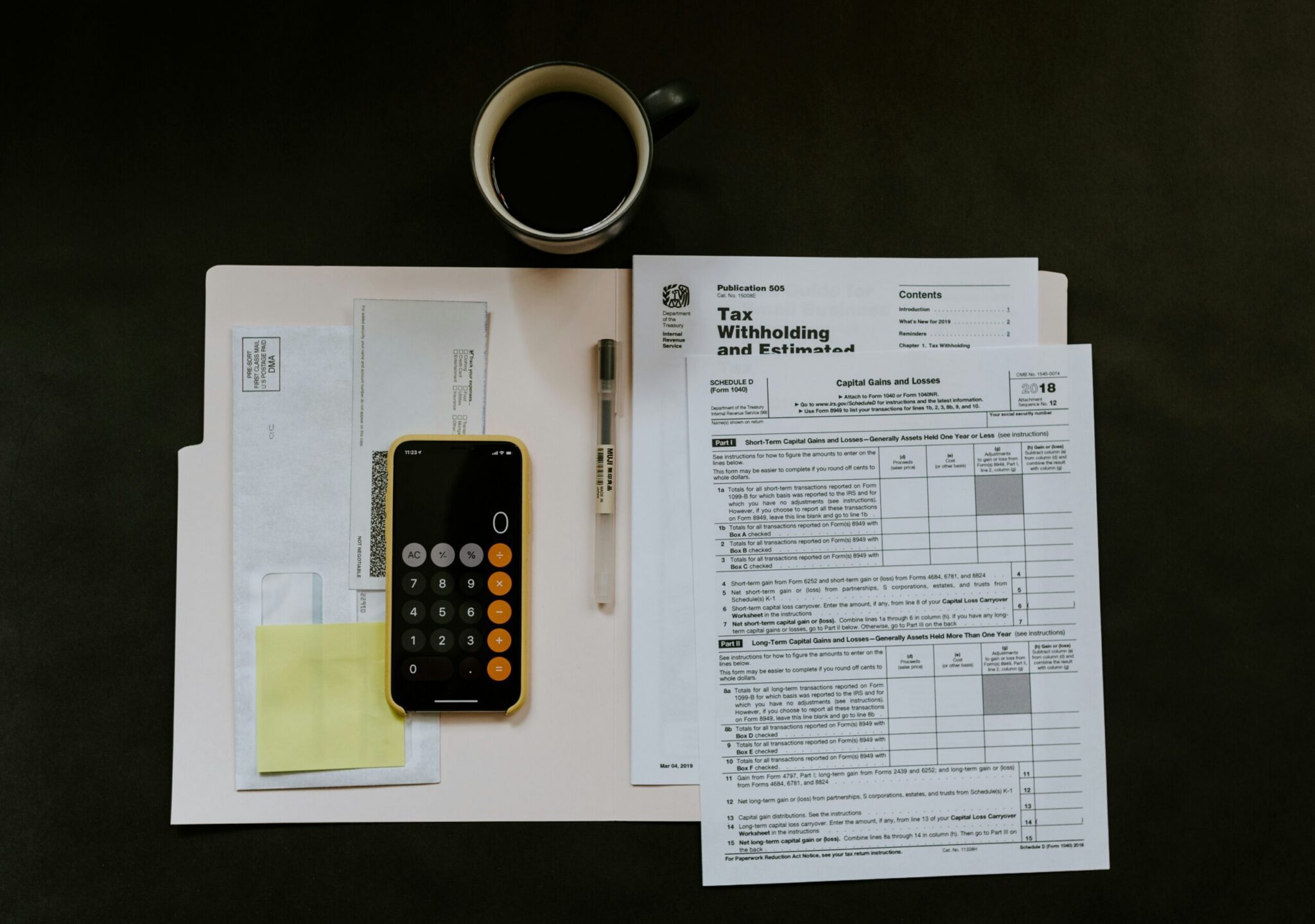 A cup of black coffee, tax forms, a smartphone with a calculator app open, a pen, an envelope, and sticky notes arranged neatly on a dark surface.