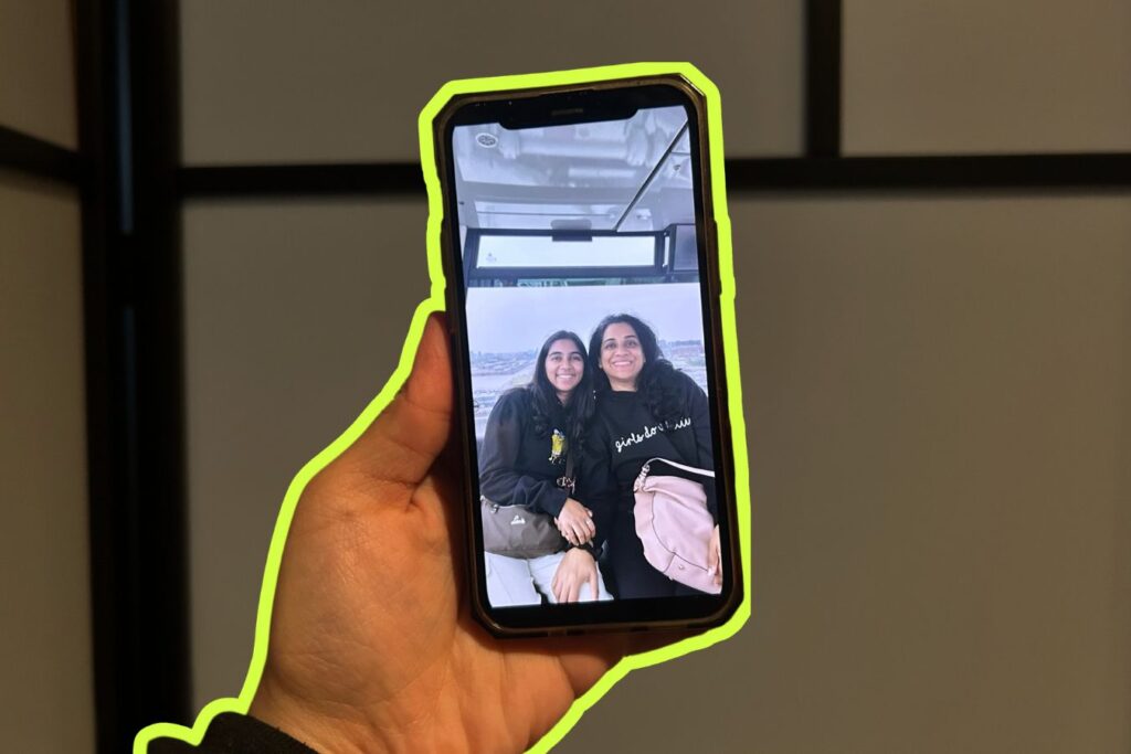 A hand holding a phone displaying a photo of two smiling women in a bus. The phone is outlined with a neon green border. The background shows an white divider with wood panels.