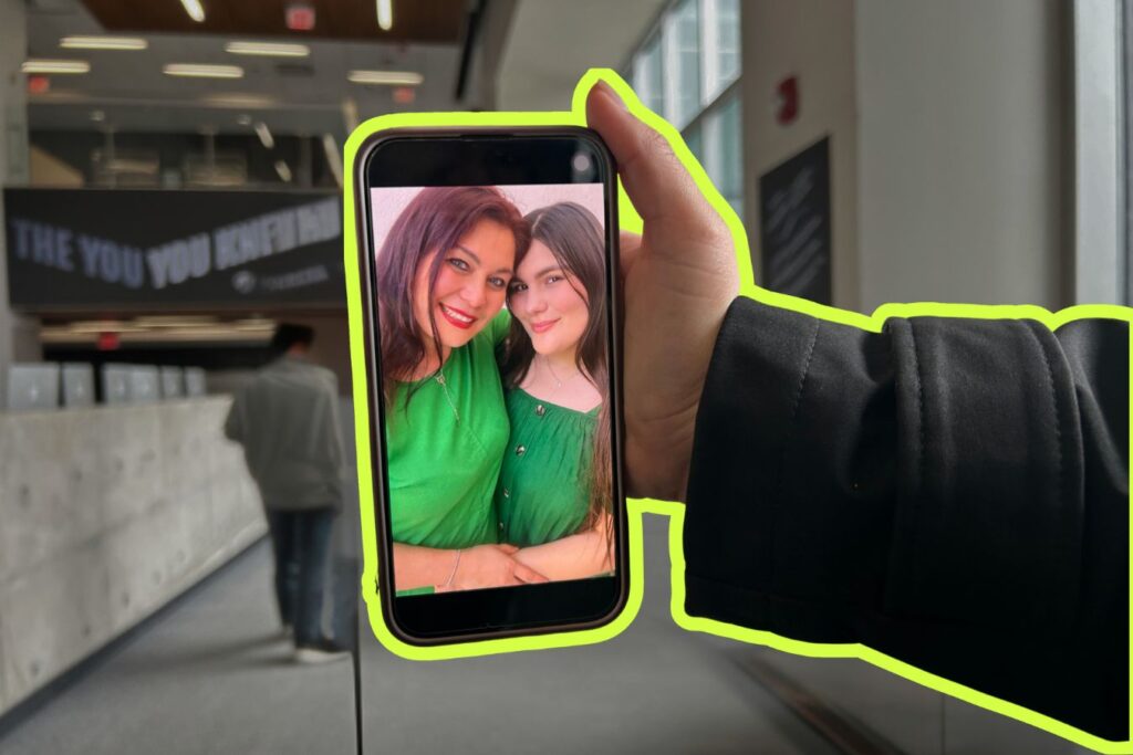 A hand holding a phone displaying a photo of two smiling women: a mom and a daughter. The phone is outlined with a neon green border. The background shows the entrance of L building at Humber Lakeshore Campus.