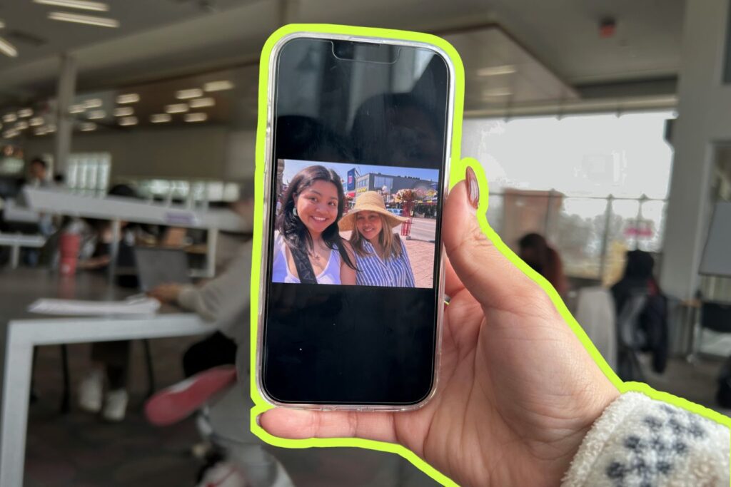 A hand holding a phone displaying a photo of two smiling women outdoors; the phone is outlined with a neon green border. The background shows an indoor space with people working at tables.