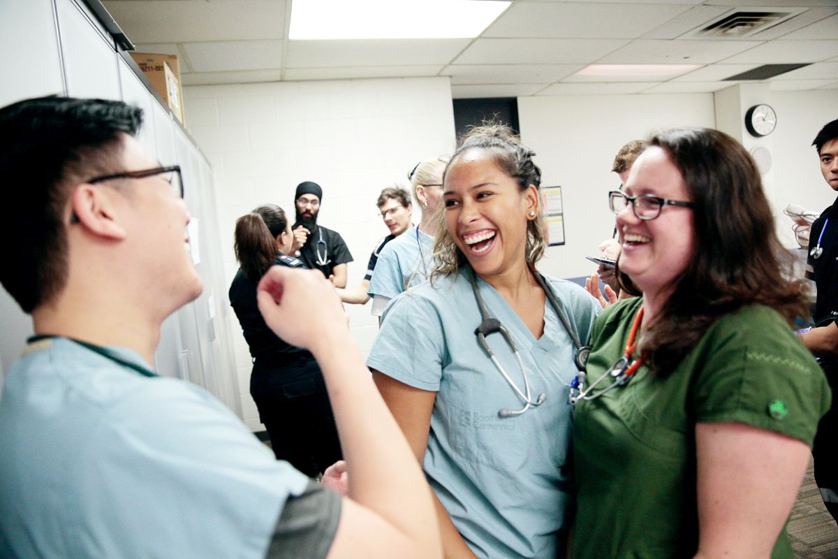 A medium, eye-level shot of a diverse group of healthcare or nursing students sharing a joyful laugh in a brightly lit classroom or clinical lab. In the center, a smiling woman with dark hair tied back, wearing light blue scrubs and a stethoscope around her neck, laughs heartily with her mouth open. To her right, a woman with glasses and wavy brown hair in dark green scrubs and a red-tubed stethoscope laughs along warmly. On the left, a young man with glasses in light blue scrubs is seen from a three-quarter back view, smiling as he speaks to them. In the background, other classmates in various scrubs—including a Sikh man with a black turban and a stethoscope—are seen conversing near a row of tall grey metal lockers against a white brick wall.