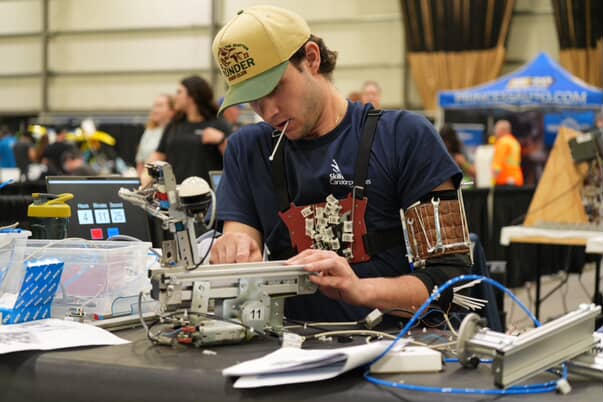 A young man wearing a baseball cap and a dark blue shirt labeled "Skills Canada" focused on assembling or adjusting a complex robotic mechanism on a workbench. He holds a zip tie in his mouth while wearing customized gear carrying tools and components across his chest and upper arm in a large event venue.