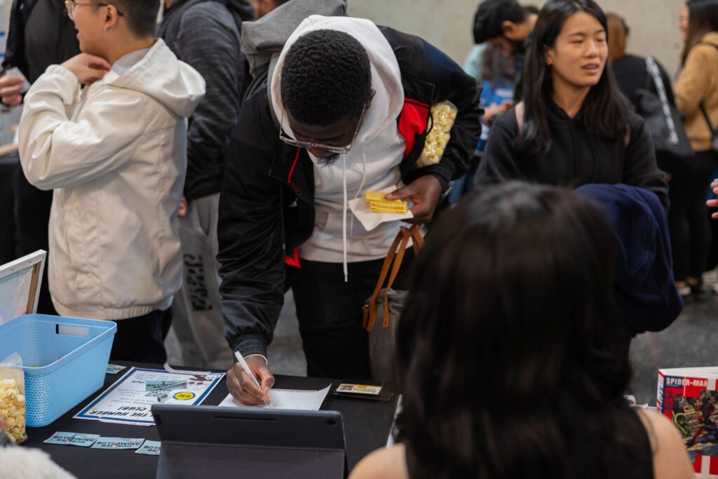 A student wearing glasses leans over a table to write on a sign-up sheet at an event.