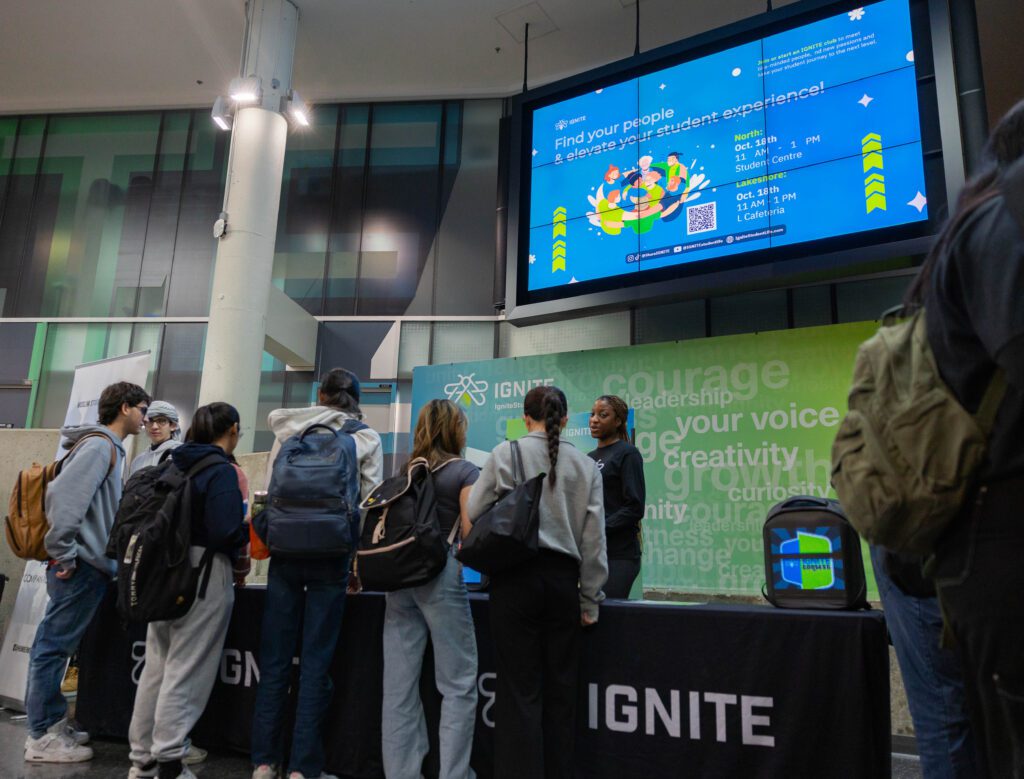 A group of students with backpacks gathers around an IGNITE booth at a student event.