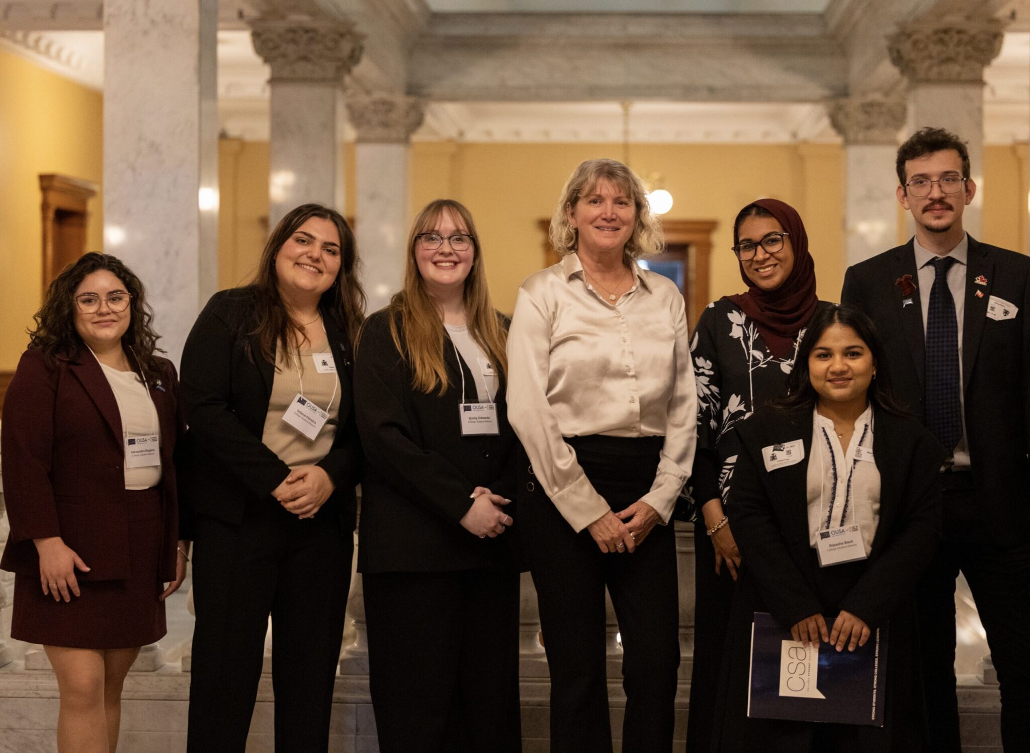 A group photo of seven participants in professional business attire posing together inside a grand hall featuring ornate marble columns during the College Student Alliance (CSA) Advocacy Summit. They stand smiling at the camera with event name badges, including six student delegates and a featured guest standing at the center.