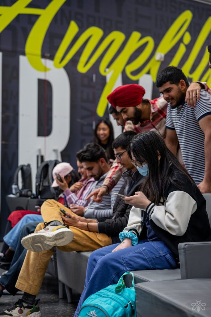 A diverse group of people are gathered indoor space with modern seating while using their smartphones and engaging with each other.