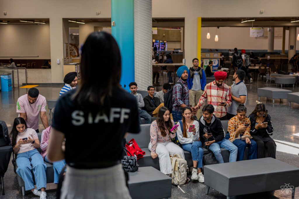 A staff member in a black shirt stands in the foreground addressing a diverse group of students.