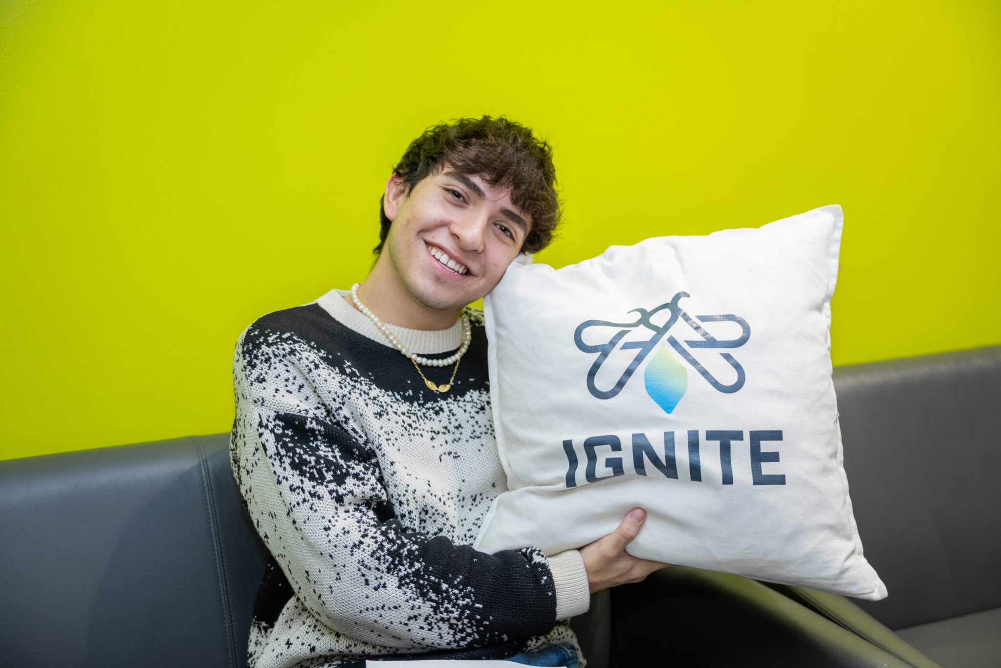 A young man wearing a black-and-white patterned knit sweater and pearl necklace sits on a grey leather sofa in the IGNITE clubs room. He smiles warmly at the camera while resting his head against a white throw pillow printed with the IGNITE logo, set against a vibrant lime green accent wall.