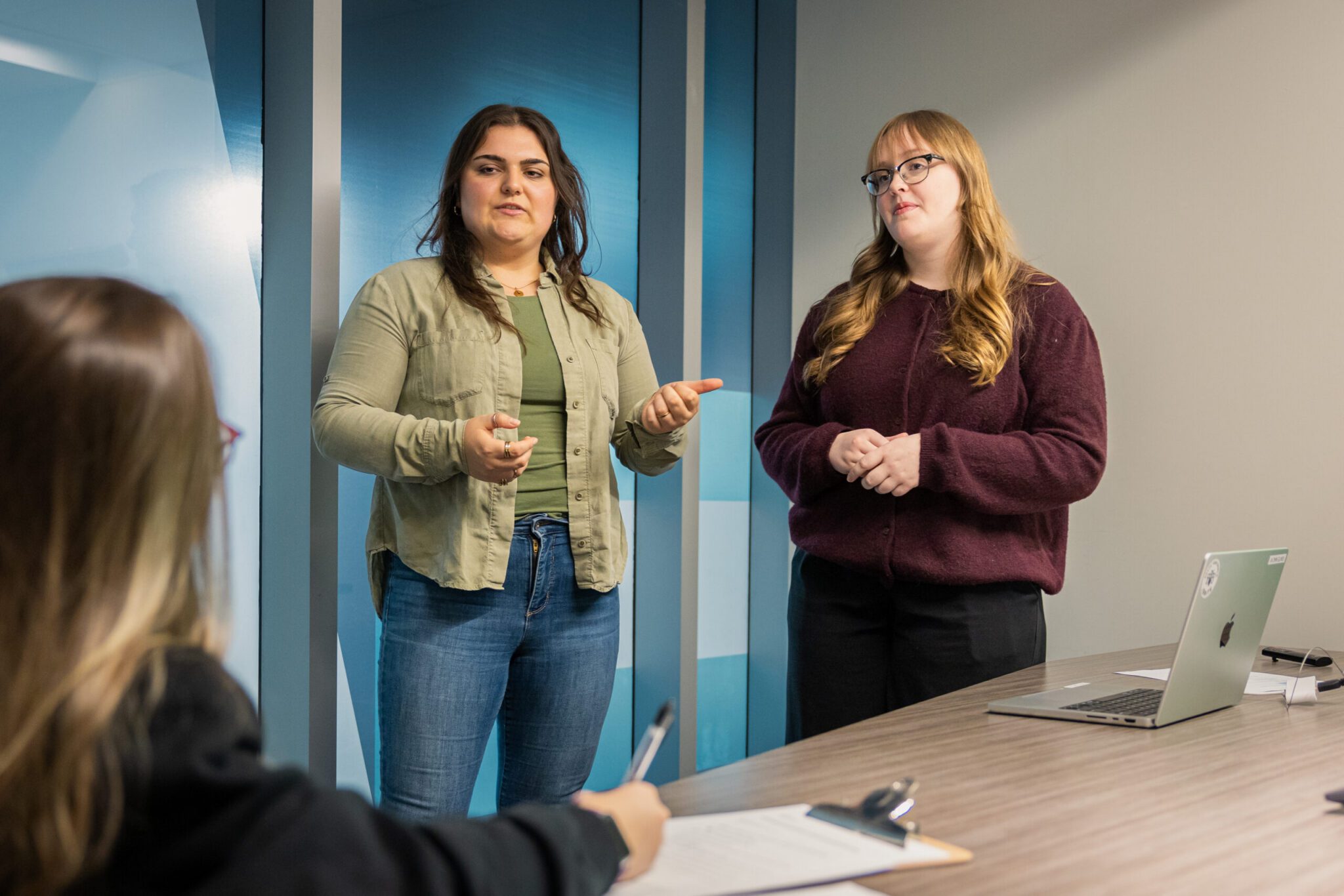 Two IGNITE Student Advocates standing at the head of a wooden conference table during a presentation. The advocate on the left, with dark hair wearing an olive green shirt over a matching tee and blue jeans, gestures with her hands as she speaks. Next to her, an advocate with blonde hair, glasses, and a burgundy sweater stands attentively with hands clasped. In the foreground, a seated person takes notes on a clipboard, with an open silver laptop on the table.