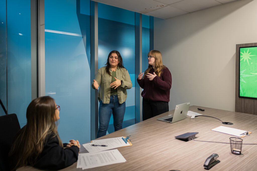 IGNITE Student Advocates are standing and gesturing while presenting in a modern conference room. A seated woman listens attentively.