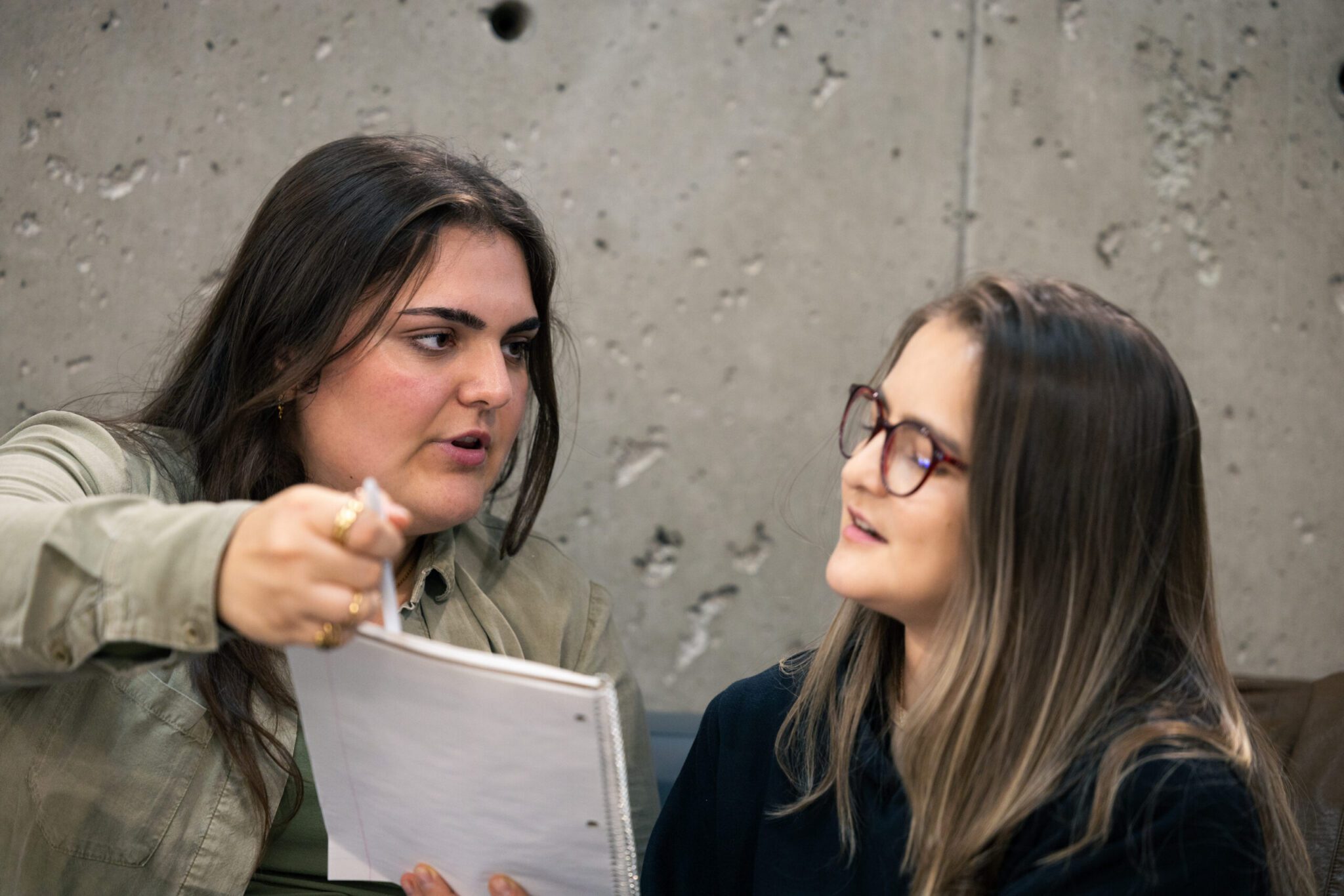 Two young women sitting together against a textured concrete wall, with one pointing to a notebook while explaining a concept to her colleague.