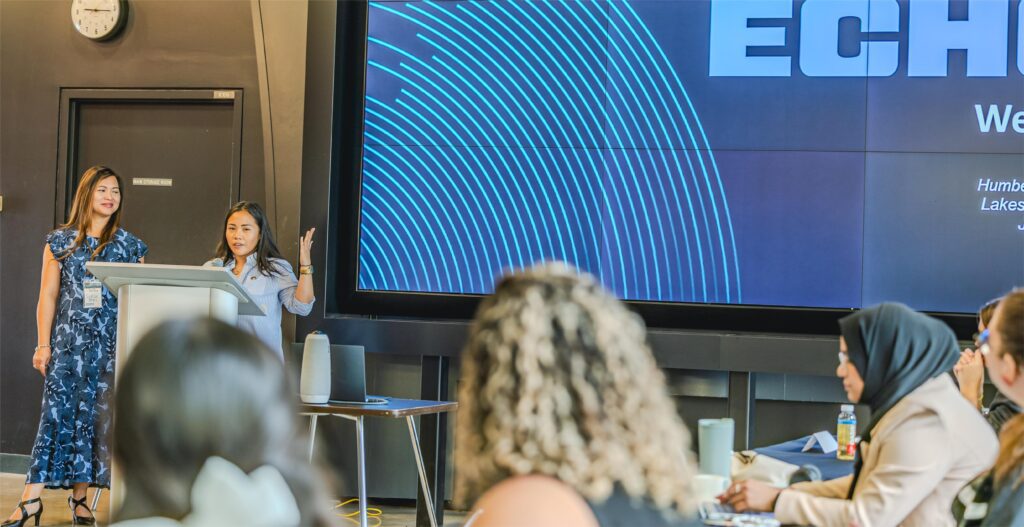 Two women are speaking at a conference and a group of people are sitting and listening.