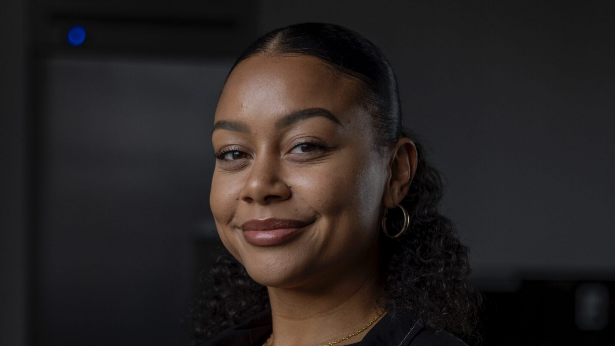 A medium portrait of a smiling woman with curly dark hair pulled back into a low ponytail. She wears a short-sleeved black button-up shirt with prominent white contrast stitching, white trousers, gold hoop earrings, and a delicate gold necklace, seated against a dark background.