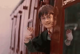 A boy with glasses on a train waving his hand as the train moves away.