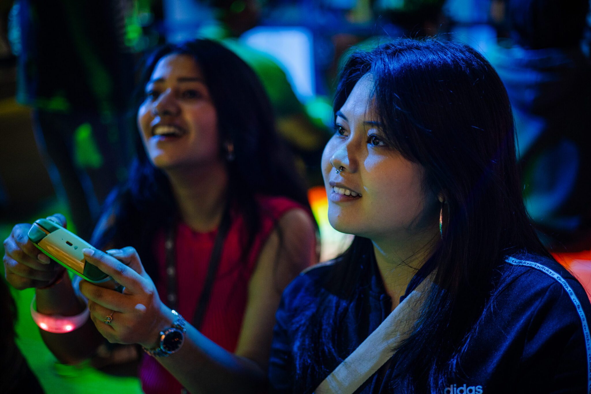 A candid shot of two young women playing video games together in a venue illuminated by blue and green neon lighting. In the foreground, a woman with dark hair and a nose piercing looks attentively toward the screen with a subtle smile. Beside her, a friend holding a video game controller laughs excitedly.