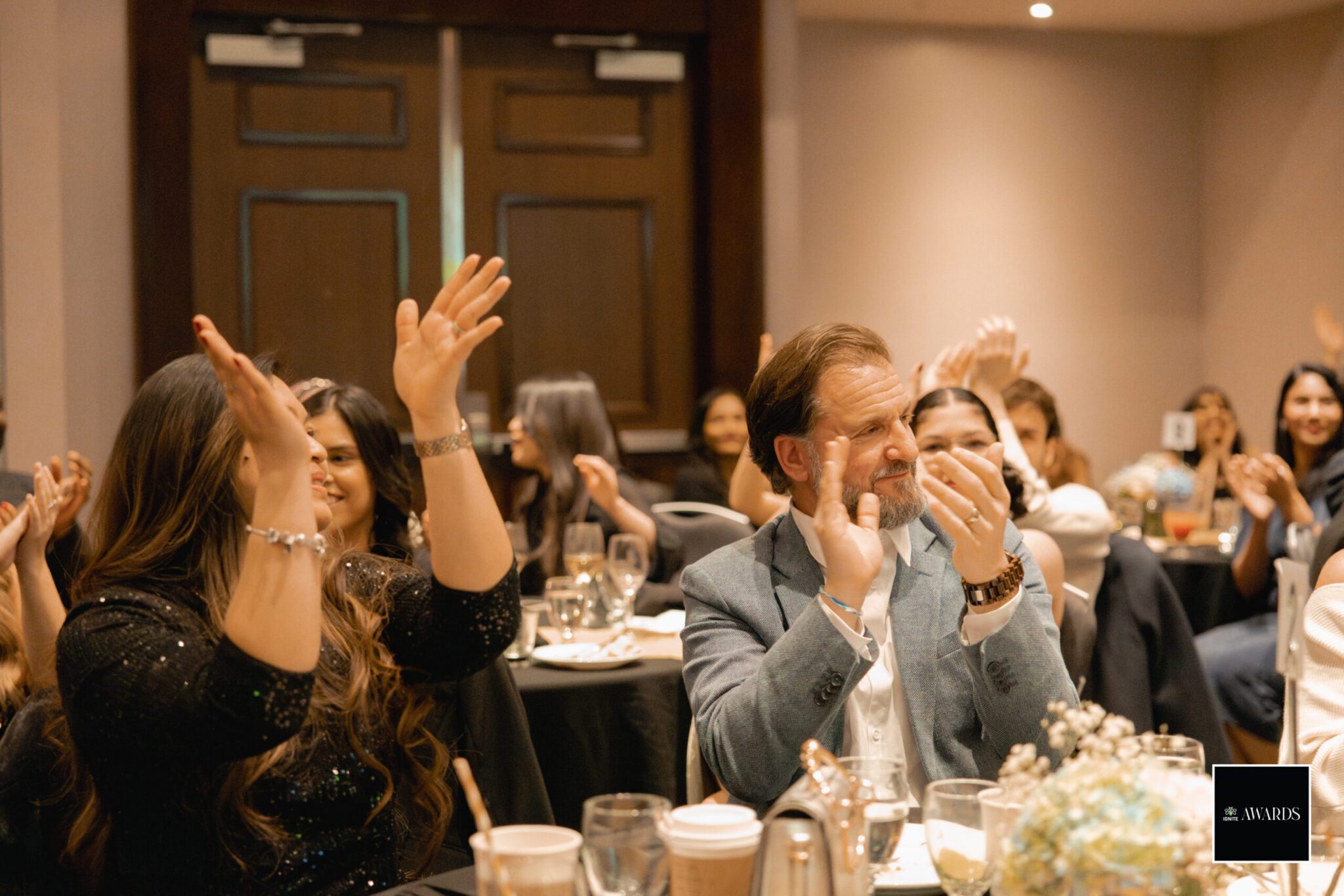 A candid shot of attendees seated at formal banquet tables applauding during the IGNITE Awards 2024 event. In the center, IGNITE CEO Ercole Perrone, wearing a light blue blazer, smiles and claps alongside a woman in a sparkly black sequin top cheering with raised hands. An "IGNITE AWARDS" logo sits in the bottom right corner.