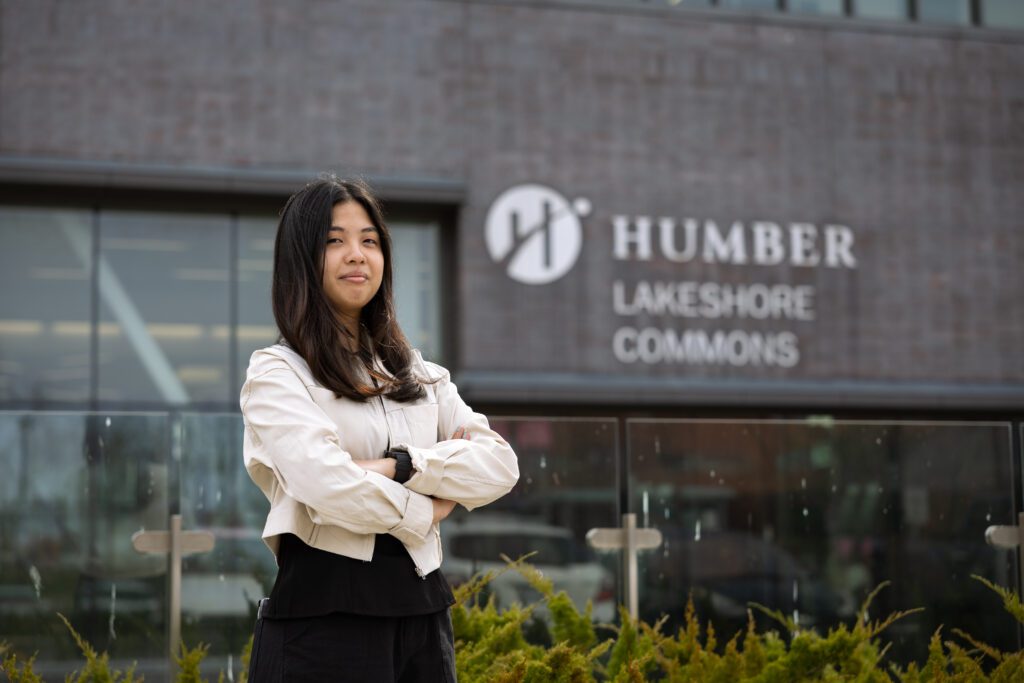 A woman is smiling while standing in front of the Humber Polytechnic Lakeshore campus. 