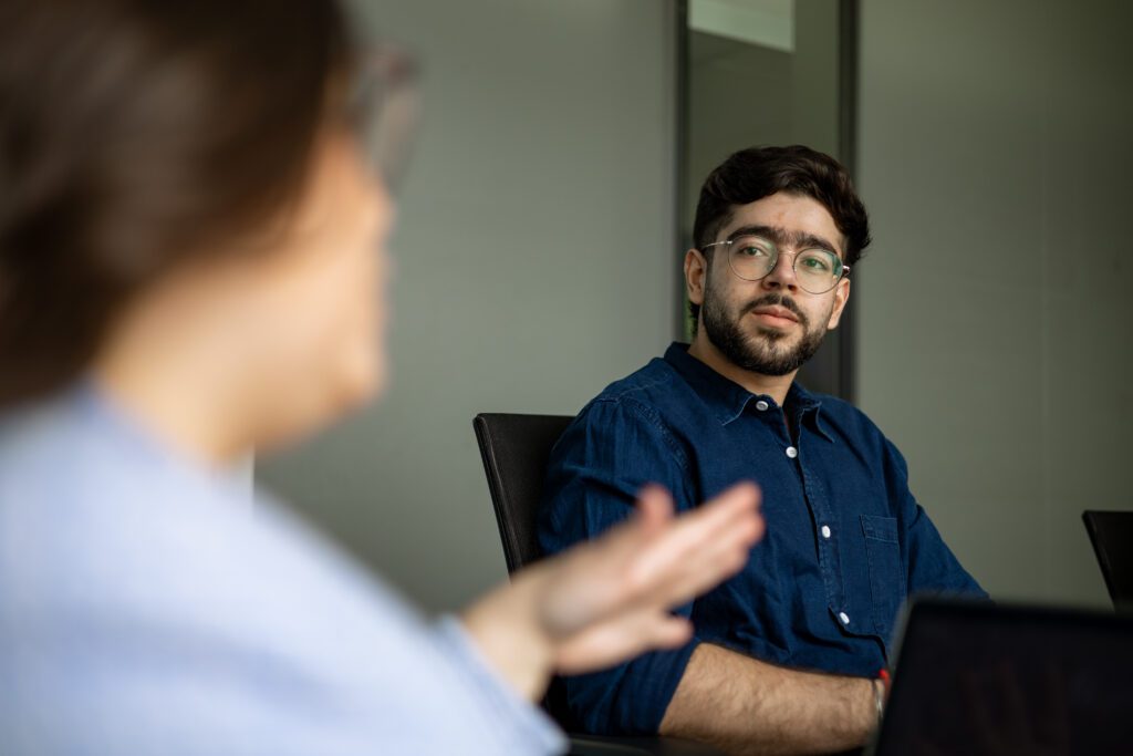 A Board of Directors member wearing glasses and a blue shirt sits and listens to a person speaking during a meeting in an office setting.