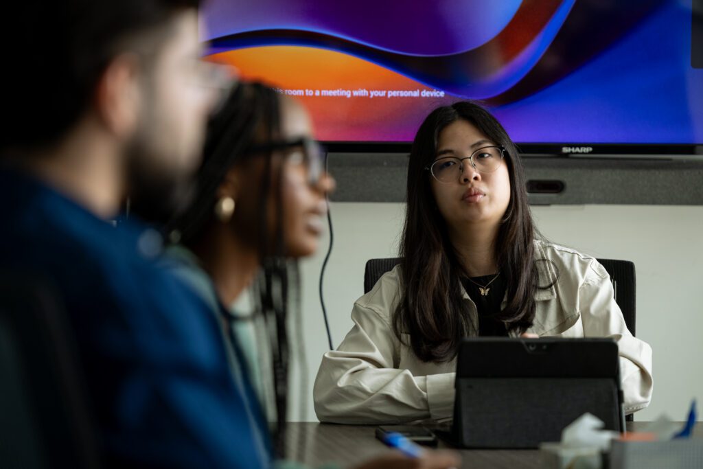 Three people are sitting at a conference table. One woman in focus and looking attentive, with a monitor with abstract colours in the background.