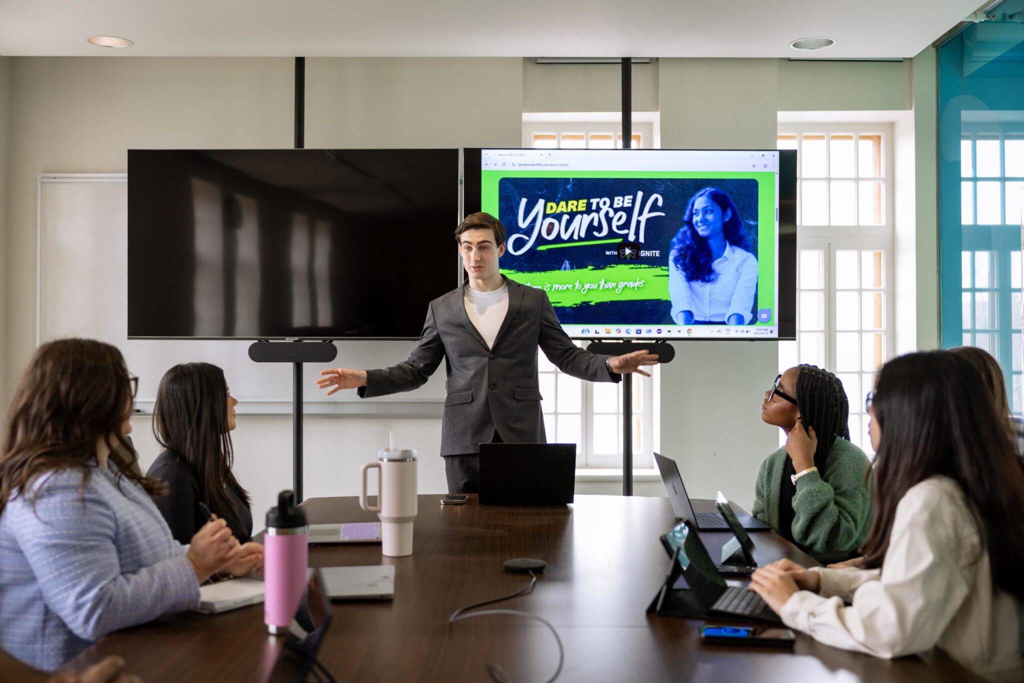 A young man in a grey suit blazer stands at the head of a conference table with open arms, presenting to a group of seated colleagues in a board room with a large screen behind him displaying IGNITE's "Dare to Be Yourself" graphics.