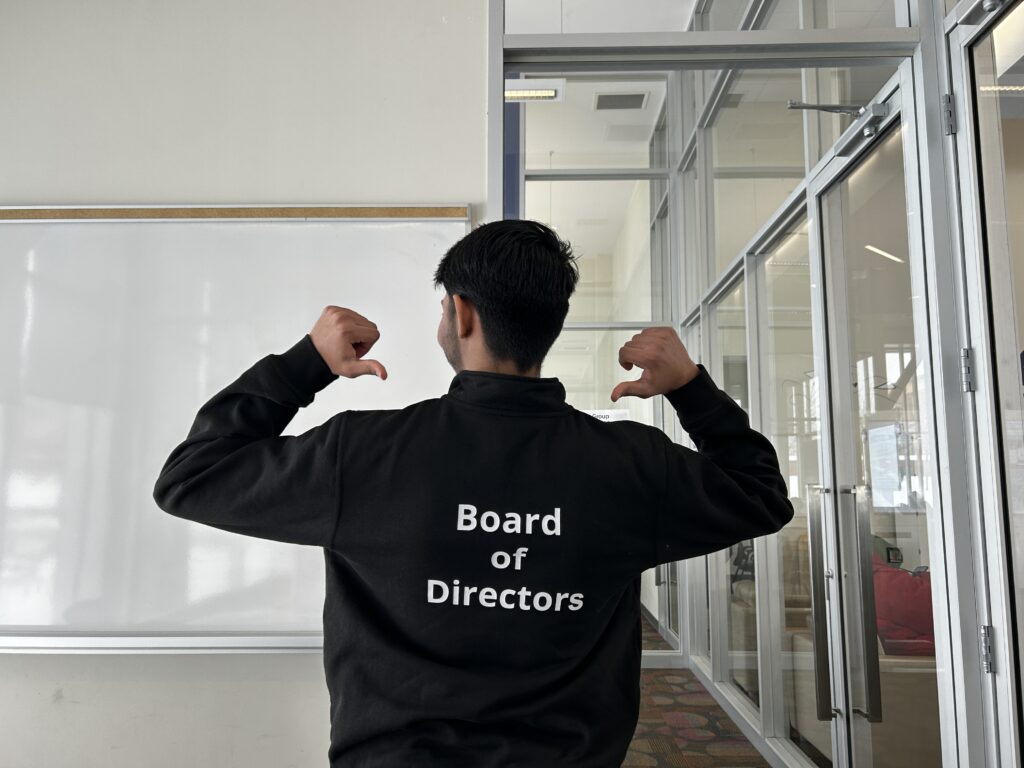 Kartavya Patel shows the text at the back of his sweater, his two thumbs pointing at the print. The text reads: "Board of Directors."
