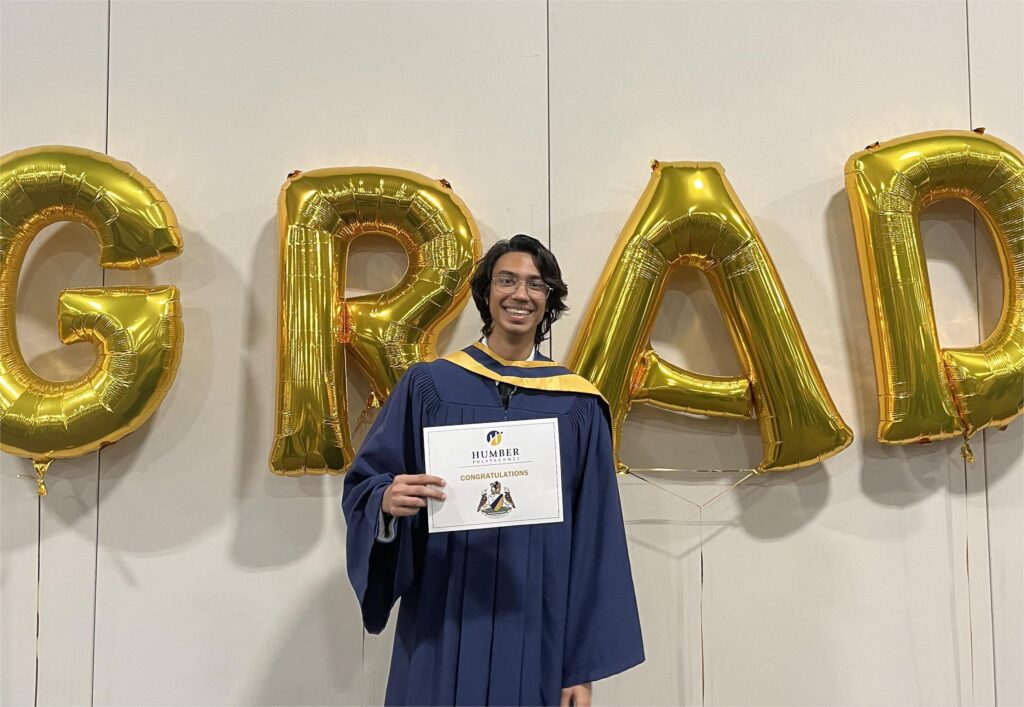A man is smiling while holding his degree certificate, with "GRAD" written in the background with balloons.