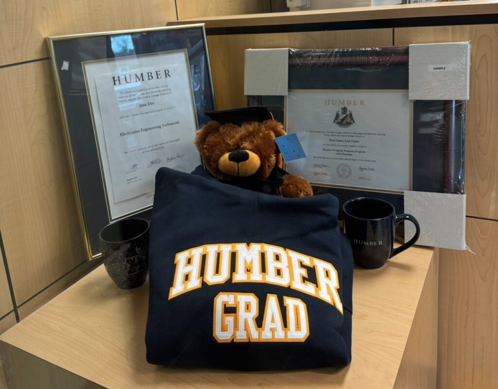 A display featuring Humber graduation memorabilia, including a black hoodie with 'HUMBER GRAD' in bold yellow and white letters, a plush teddy bear wearing a graduation cap, two black Humber-branded mugs, and two framed certificates.
