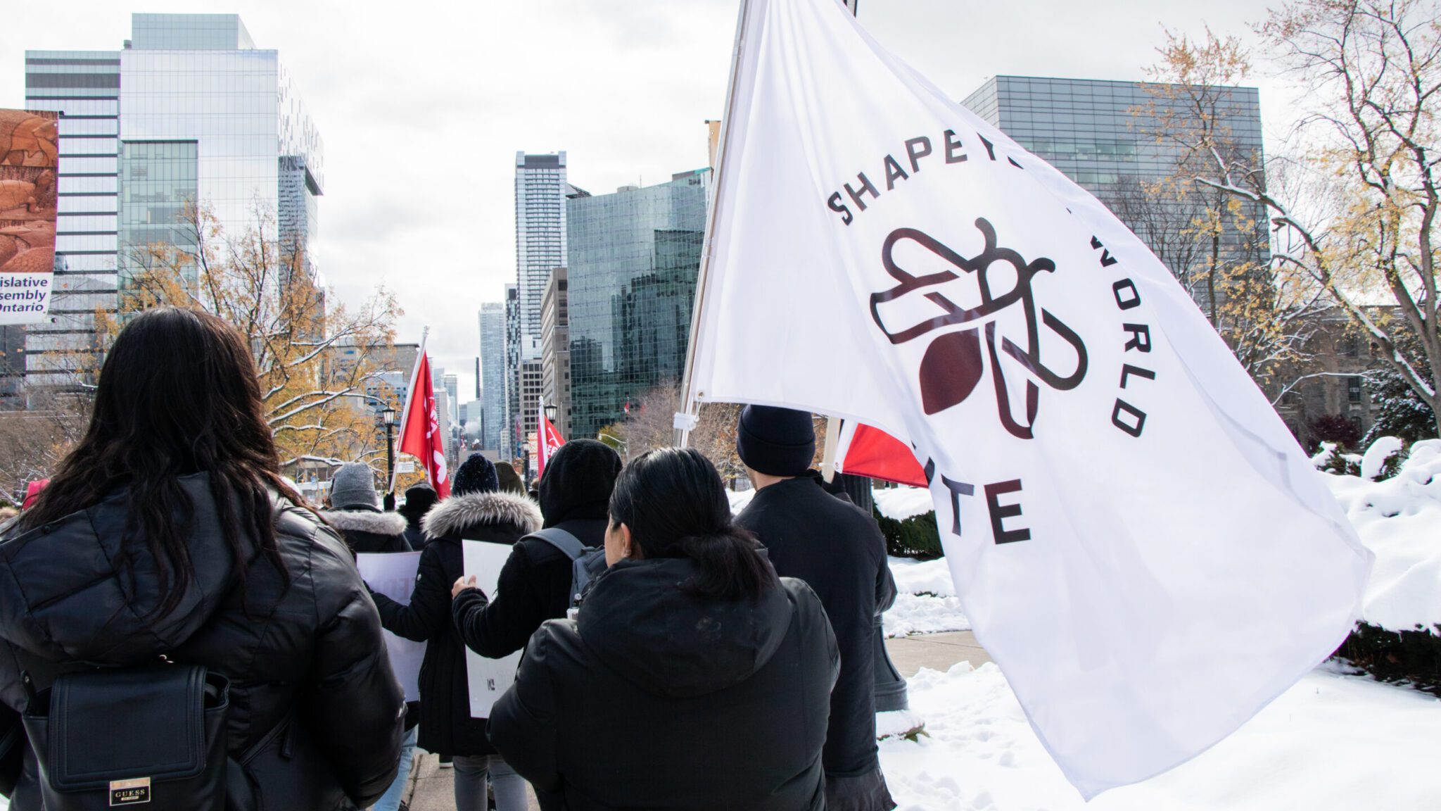 A perspective shot from behind a crowd marching along a snowy sidewalk near the Legislative Assembly of Ontario. In the foreground on the right, a large white flag featuring the brown IGNITE logo and text "SHAPE YOUR WORLD" waves over protesters dressed in dark winter coats, with modern city skyscrapers standing under an overcast sky in the background.