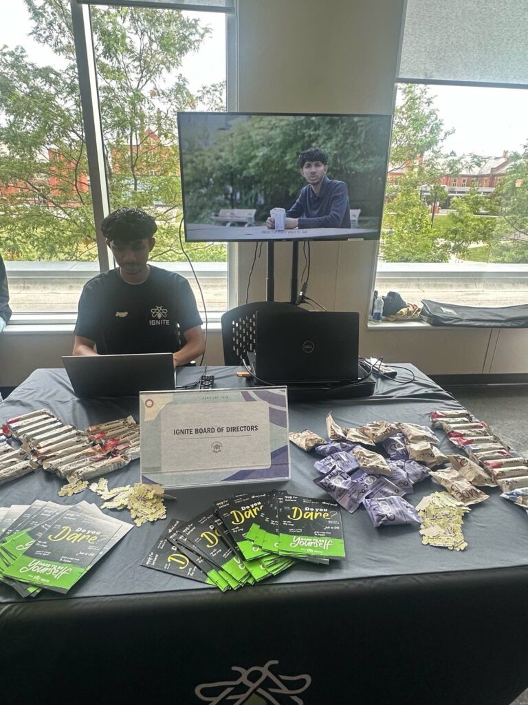 Kartavya Patel sits behind a table while using his laptop. The table has a name card that reads “Ignite Board of Directors”, snacks, and IGNITE cards. Behind is a big TV screen with a video playing.