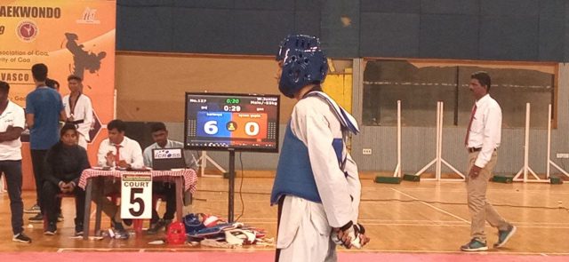 A taekwondo match in progress with one athlete, in his side profile, wears a blue protective gear standing in the middle.