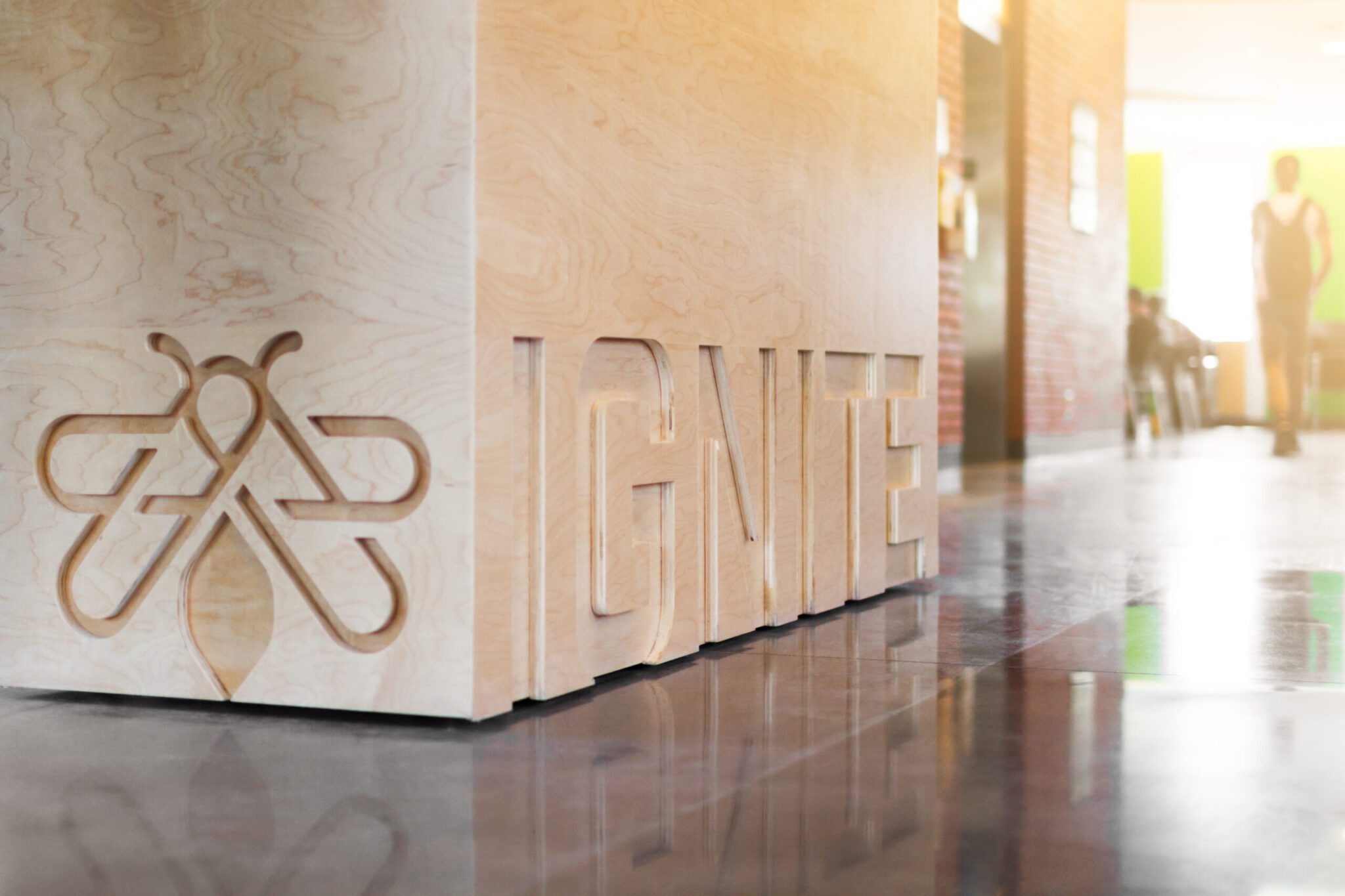 A low-angle shot of a light-colored wooden counter structure in a sunlit hallway. The front left corner features a detailed carved bee-shaped logo, while the side face displays raised wooden block lettering spelling out "IGNITE." Warm sunlight streams from a doorway in the background, creating bright highlights and reflections along the polished floor as a blurred student walks by.