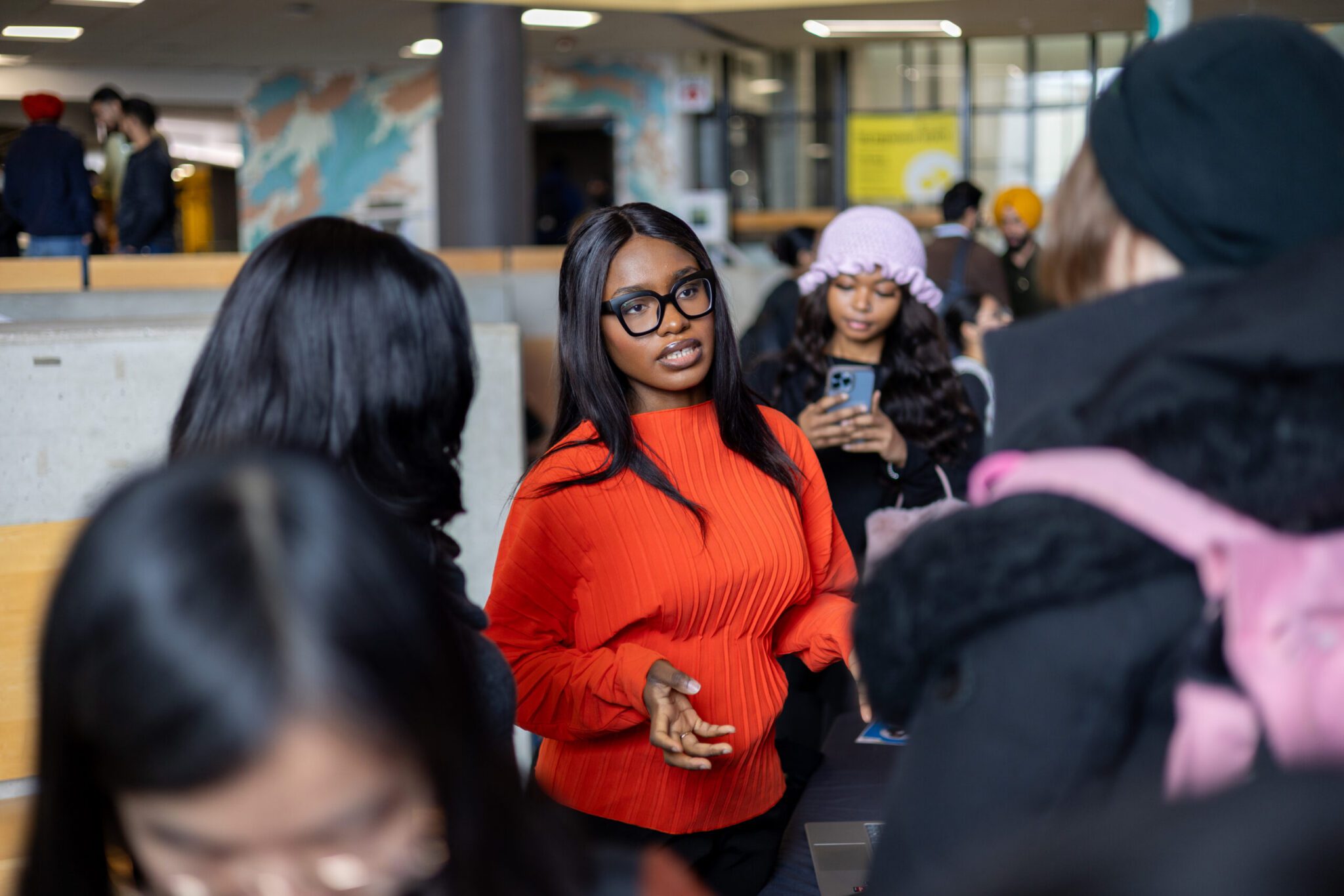 A medium shot of a young Black woman with long, straight dark hair and thick black-rimmed glasses speaking to a group. She is wearing a bright, pleated orange-red long-sleeve top and gestures with her right hand as she talks. In the blurred foreground, the backs of several students listening to her are visible. Just behind her, another student wearing a pink ruffled knit hat holds up a light-blue smartphone, while other students, including one wearing a yellow turban, stand in a spacious campus lobby with concrete pillars and a colorful wall mural in the background.