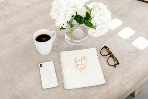 A neutral-toned workspace featuring a cup of black coffee, a white smartphone, tortoiseshell glasses, and a vase of white hydrangeas on a textured table.