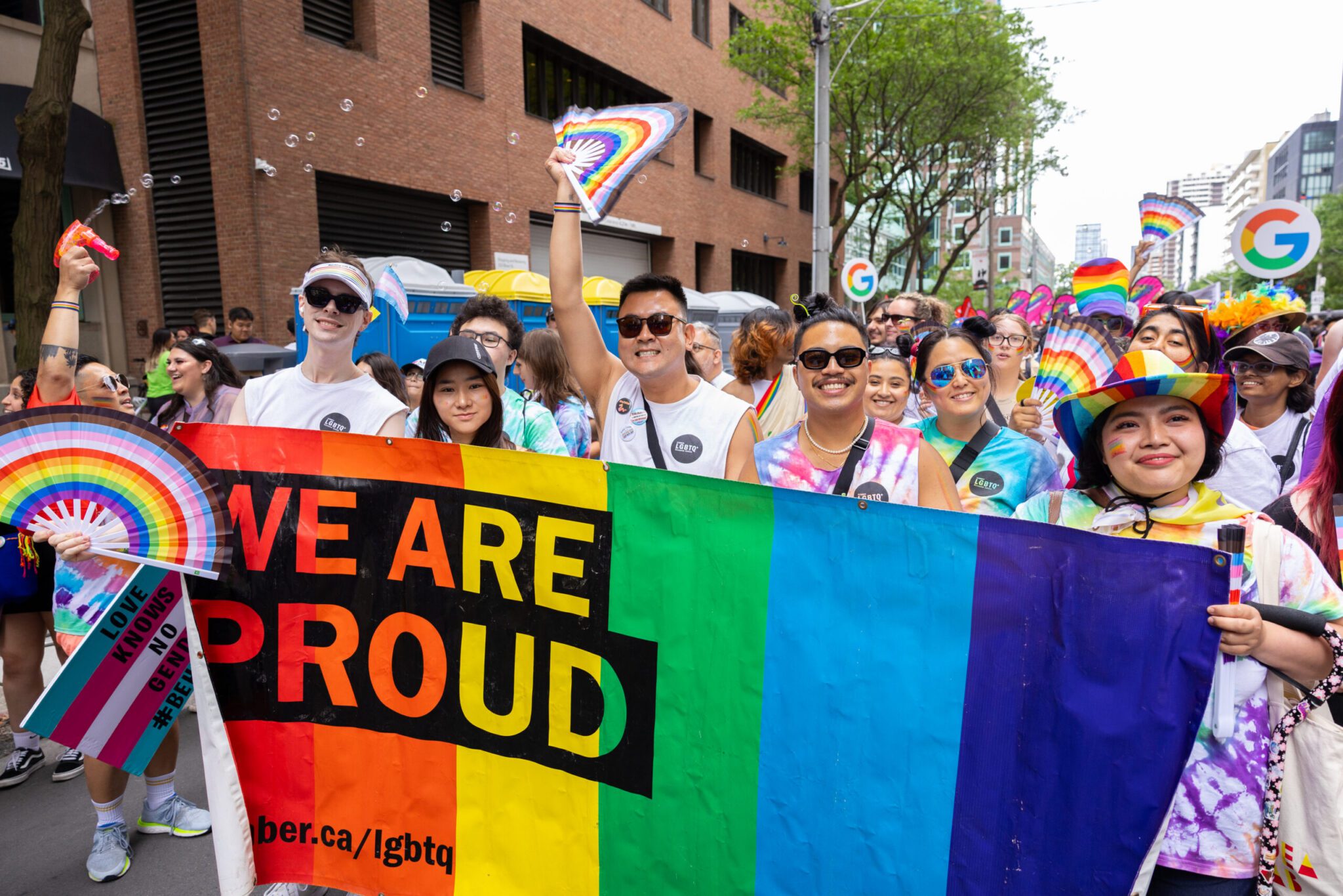 A lively photo of a diverse group of smiling participants marching outdoors in a Pride parade while holding a large rainbow banner that reads "WE ARE PROUD." Many people in the crowd wear colorful tie-dye shirts, sunglasses, and festive hats while fanning themselves with rainbow folding fans as soap bubbles float around them in a city street setting.