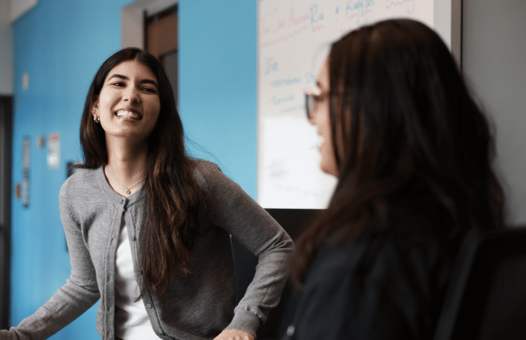 Two young women are engaged in a joyful conversation, with one smiling brightly in a gray cardigan and the other, slightly out of focus, wearing glasses and a black outfit.