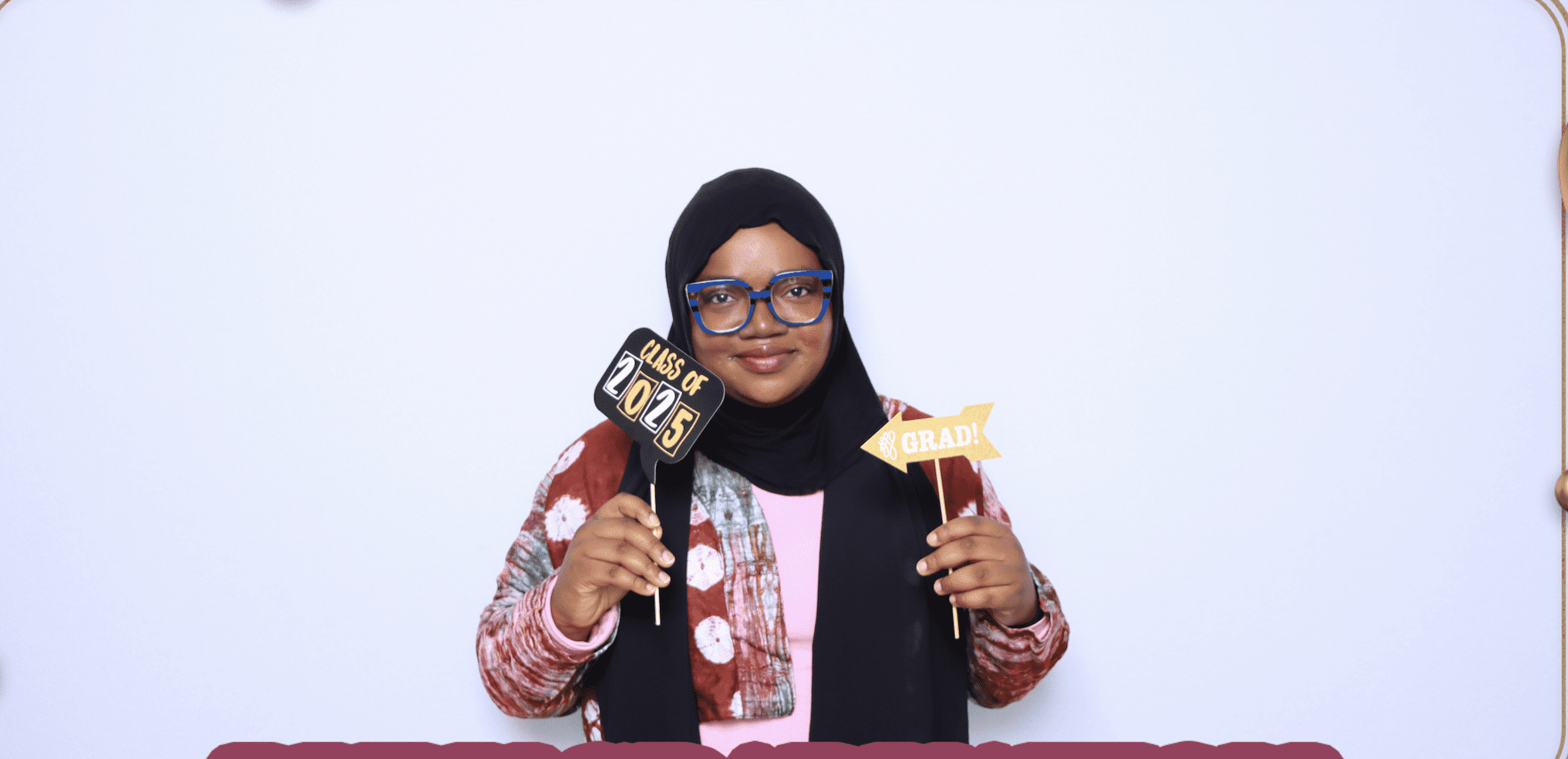 A smiling woman wearing a black hijab, blue-framed glasses, and a patterned rust-colored cardigan standing in front of a plain white studio backdrop. She holds two photo booth props on sticks: a black sign reading "CLASS OF 2025" in her right hand, and a gold arrow sign reading "GRAD!" in her left.