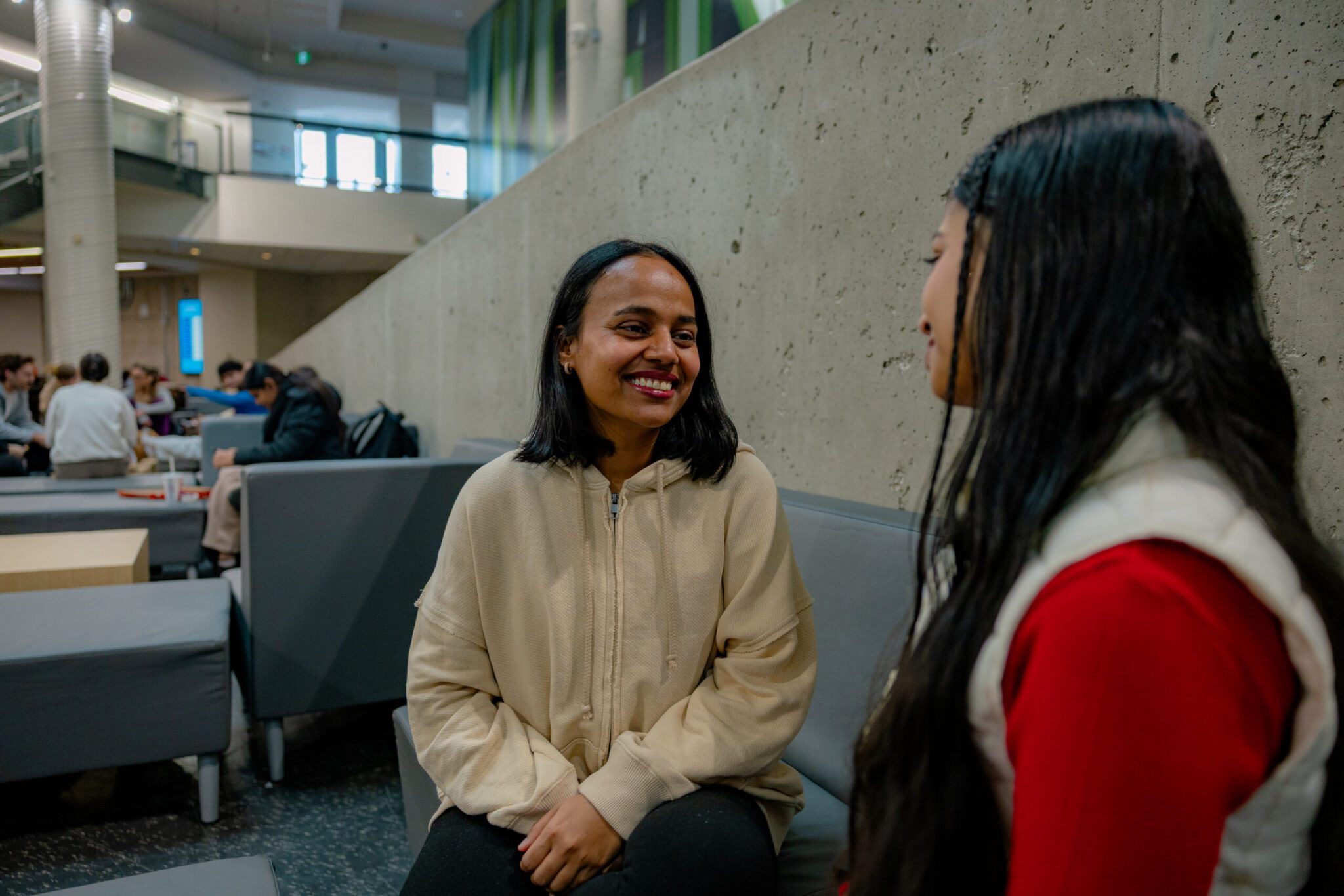 A medium shot of two young women sitting and chatting on grey modular seating inside a spacious campus student center. The woman on the left, wearing a cream-colored zip-up hoodie and dark pants, smiles warmly as she looks at her friend. The woman on the right is seen in profile with long braided dark hair, wearing a bright red top under a cream vest. Behind them, a sloping concrete wall separates the seating area from a multi-story atrium where other students are lounging and studying in the background.
