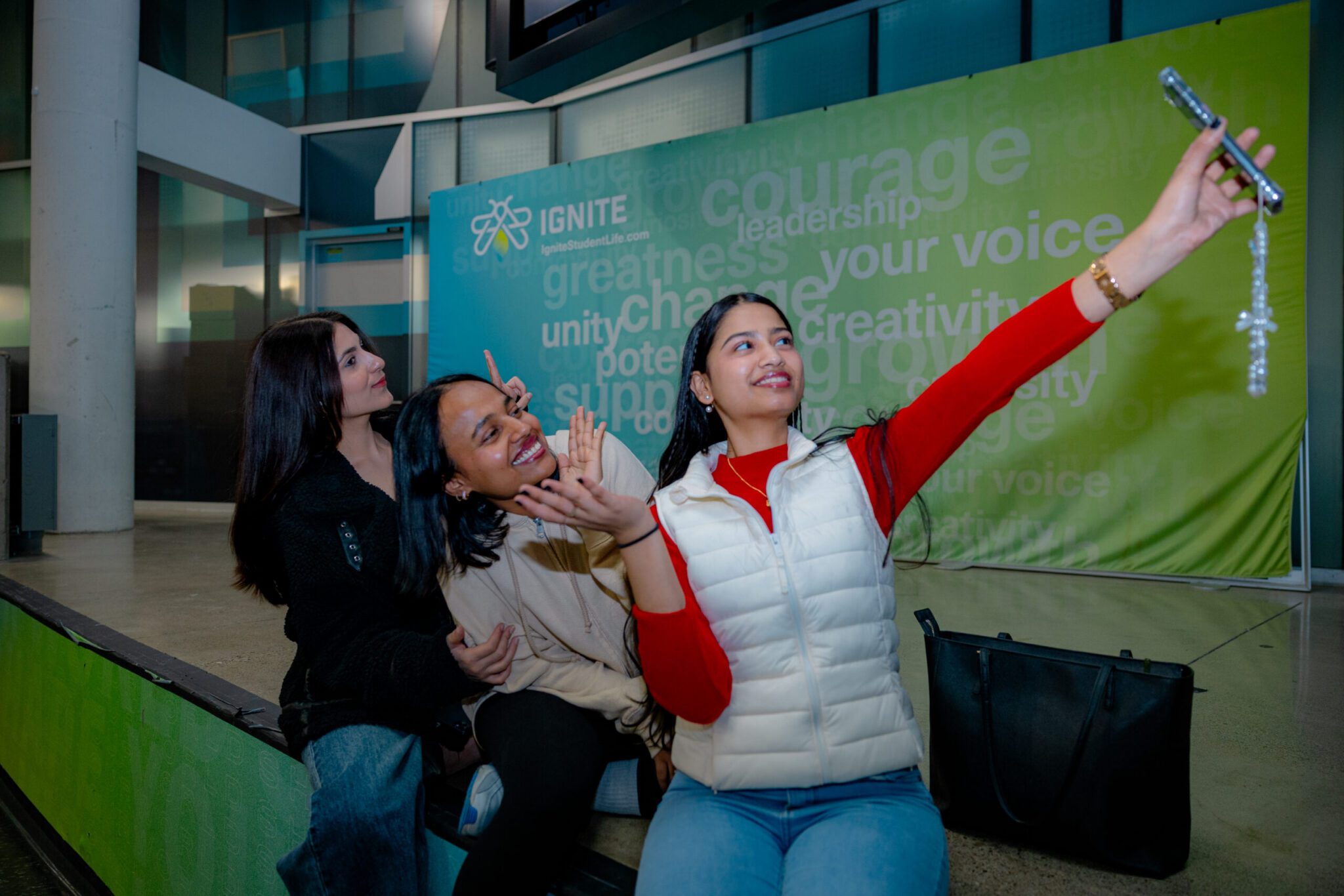 A medium shot of three young women sitting together and smiling playfully as they take a group selfie in a campus student center. The woman on the right extends her arm upward, holding a smartphone with a decorative phone charm to capture the photo, wearing a red top beneath a white puffy vest. Her two friends lean in closely behind her, posing with cheerful smiles and hand gestures. Behind them stands a large green banner featuring the "IGNITE" logo alongside inspirational words like "courage," "leadership," and "your voice." A black tote bag rests on the floor beside them.