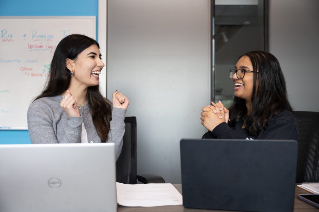 Two women sit at a table with laptops, smiling and gesturing excitedly toward each other. A whiteboard with colorful writing is visible in the background.