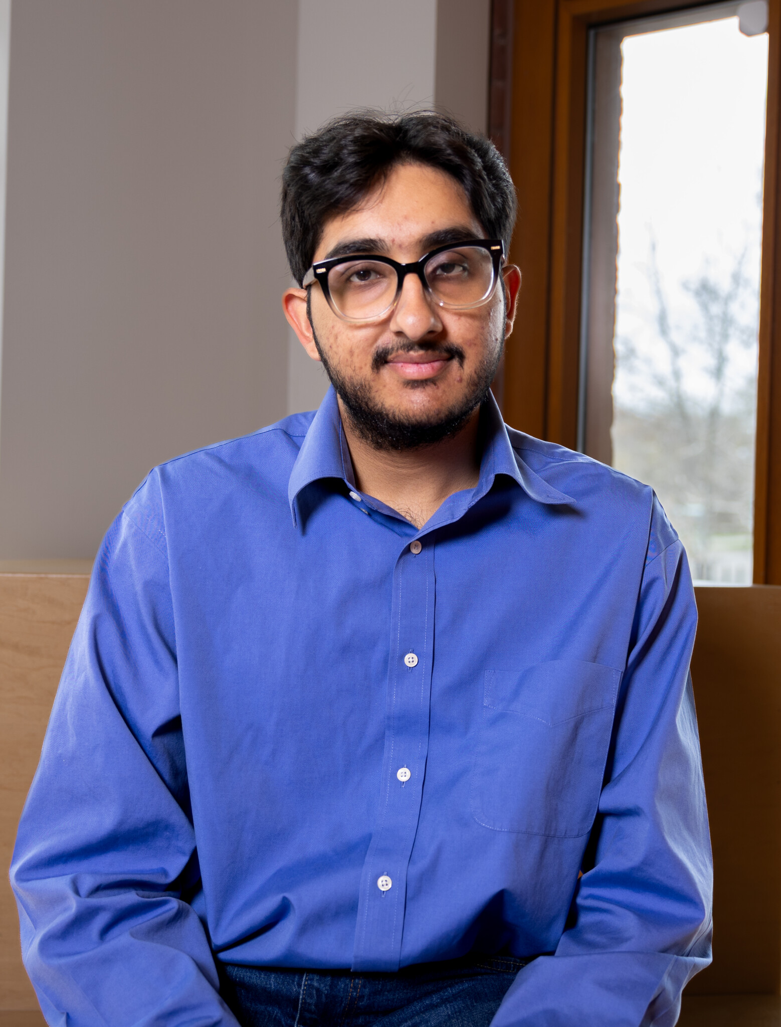 A man with glasses, dark hair, and a trimmed beard sits indoors by a window, wearing a blue button-up shirt and jeans, looking at the camera with a slight smile.