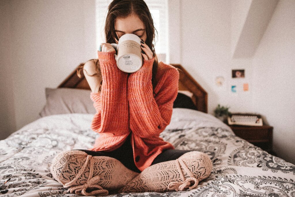A woman in a long sleeve sweater sipping a mug with both hands while in bed.