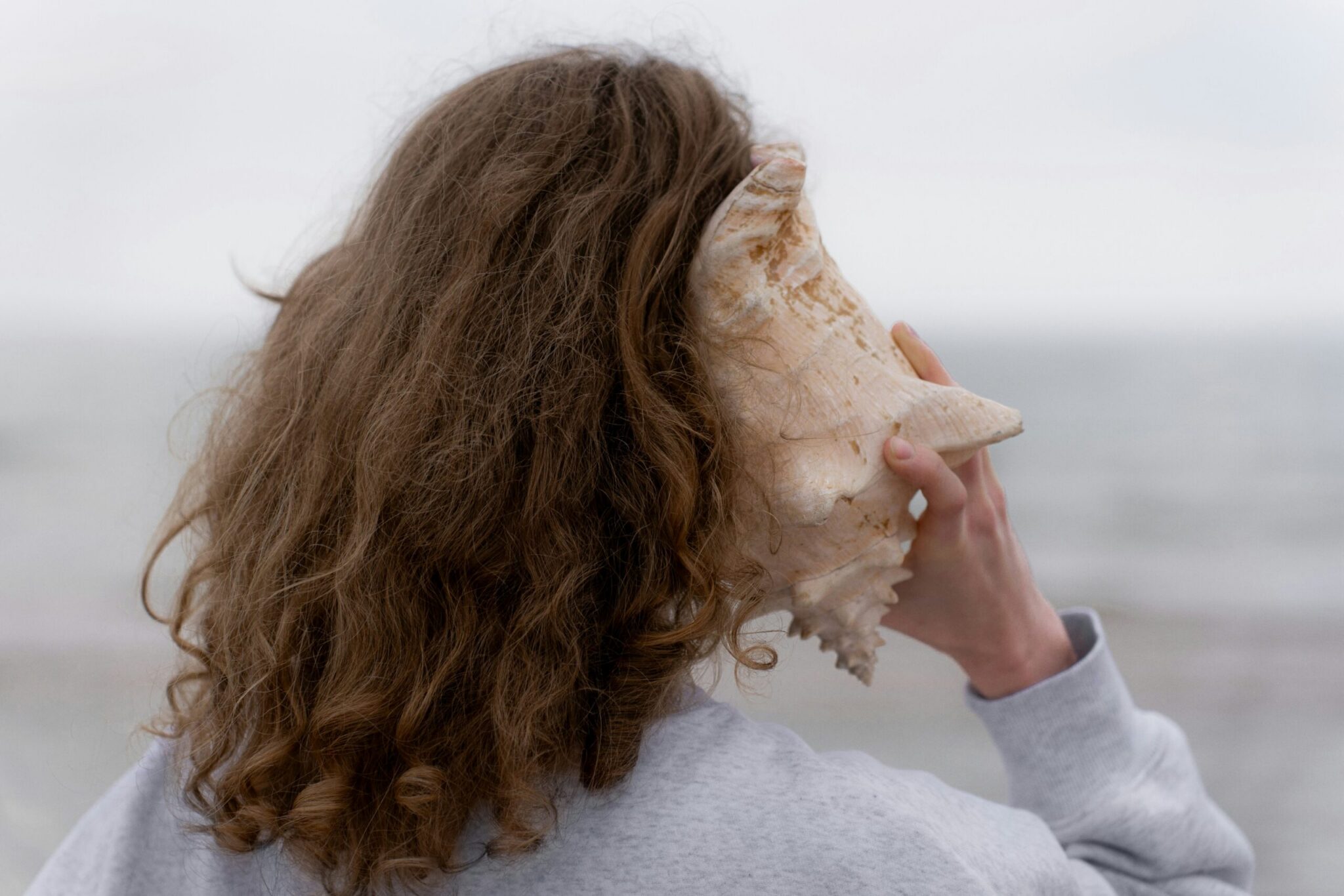 A close-up shot from behind of a woman with wavy, shoulder-length light brown hair wearing a light grey sweatshirt. She is holding a large, textured beige and pale-pink conch shell up to her right ear, as if listening to the sound of the ocean. The background shows a softly blurred, minimalist view of a calm, grey sea under an overcast sky.