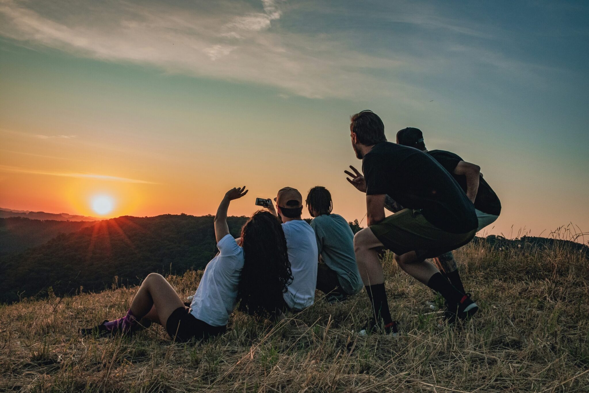 A group of five friends gathered on a grassy hilltop, facing away from the camera to watch the vibrant sun set over distant rolling hills. One person sits waving a hand while taking a smartphone photo, and another crouches making a peace sign toward the horizon.