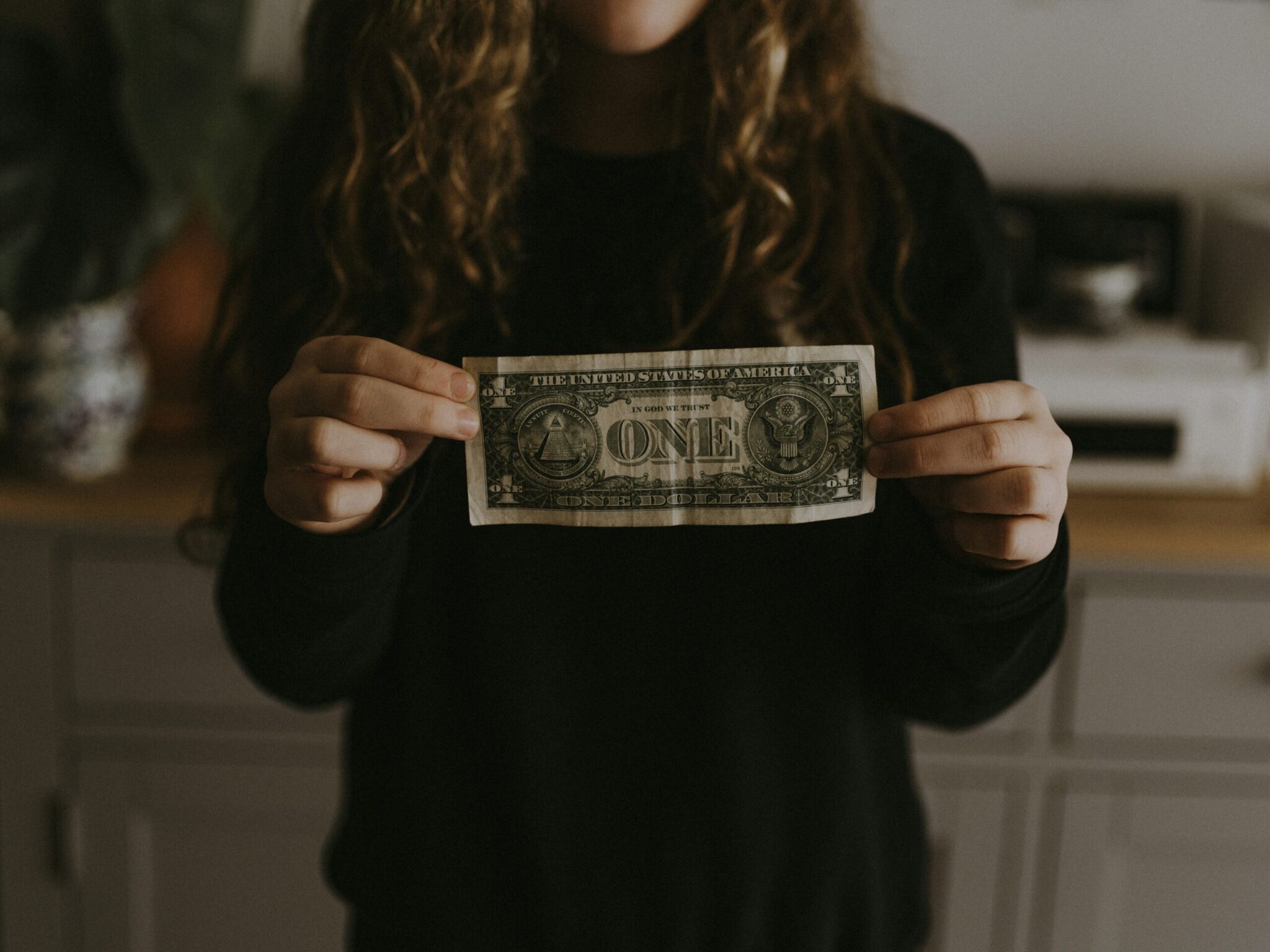 A person with long curly hair wearing a dark long-sleeve sweater holds up a US one dollar bill horizontally with both hands in front of their chest in a warmly lit indoor setting.