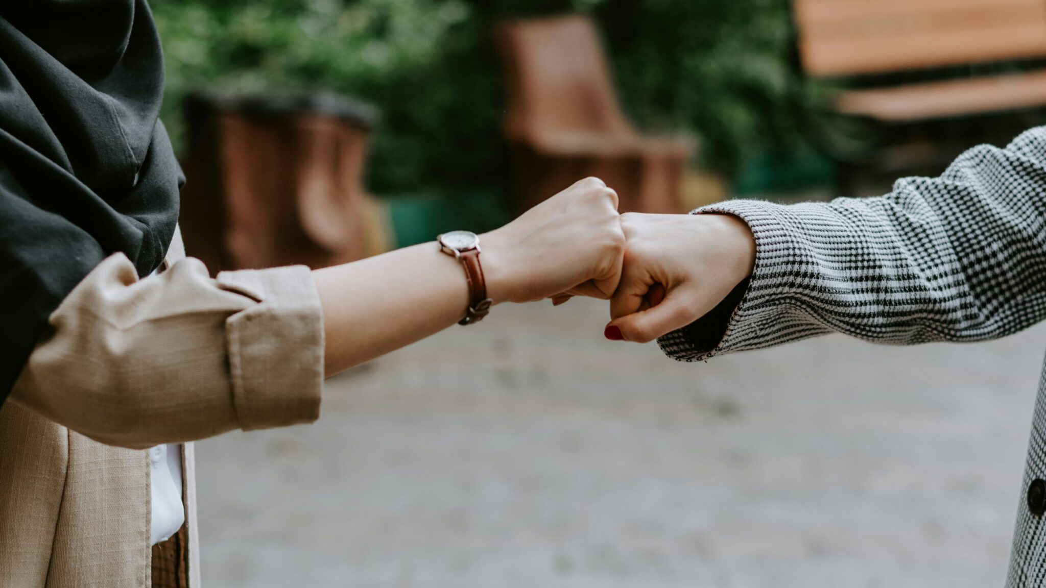 A close-up shot of two people greeting each other with a fist bump in an outdoor setting. On the left, a person wearing a black hijab, tan jacket, and wristwatch extends a fist toward another person on the right who is wearing a black-and-white patterned coat and red nail polish. The blurred background shows green foliage and wooden park benches.